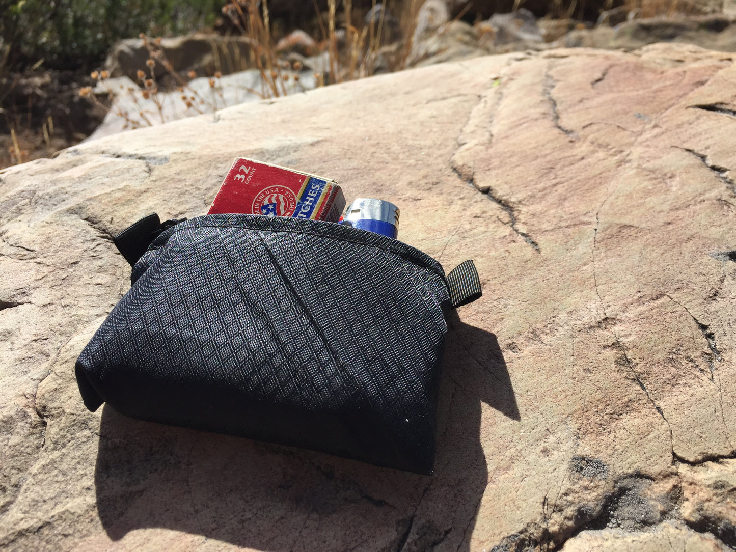 Size Small zipper pouch shown in black color option, on a rock with lighter and small box of matches